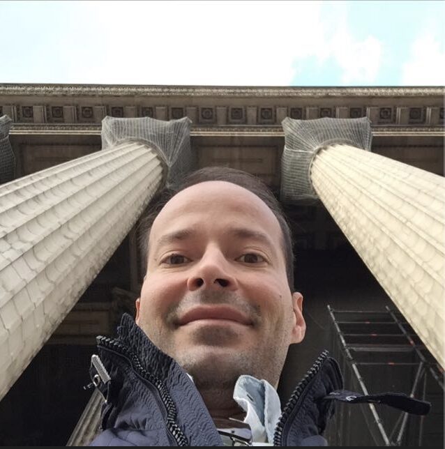 Hombre sonriendo en un selfie con grandes columnas de piedra de un edificio arquitectónico clásico en el fondo.